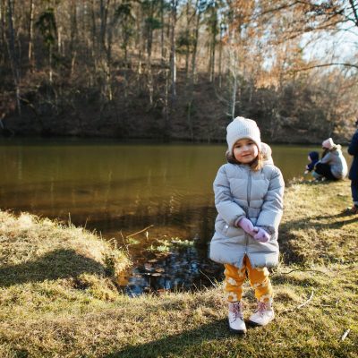 Girl in jacket stand in sunny spring park against river with family.